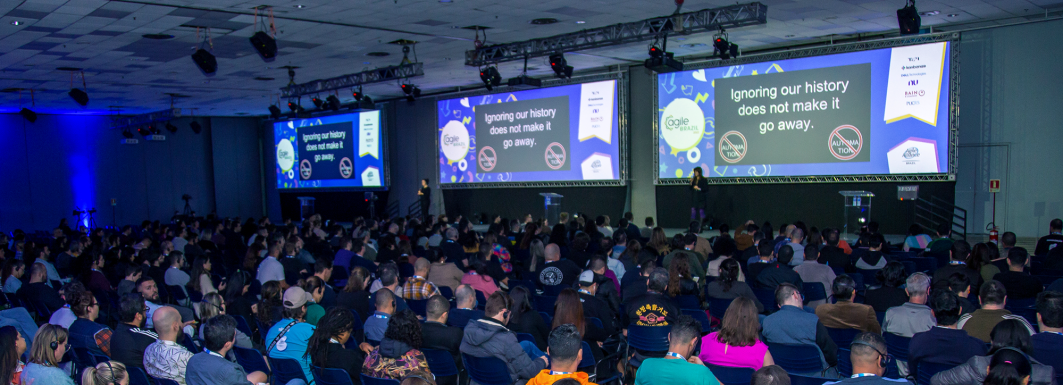 Foto de uma palestra do evento Agile Brazil. Nela tem várias pessoas sentadas de costas para a câmera e em frente às pessoas temos 3 telões com o conteúdo do palestrante.